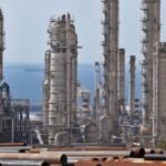 Kilang minyak dan fasilitas industri di tepi dengan latar kapal tanker di laut — jalur penting distribusi energi global melalui Selat Hormuz.