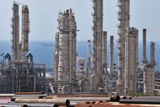 Kilang minyak dan fasilitas industri di tepi dengan latar kapal tanker di laut — jalur penting distribusi energi global melalui Selat Hormuz.