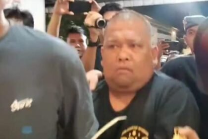 Samsul Tarigan saat digiring petugas, dikerumuni wartawan dan aparat keamanan. Meski Mahkamah Agung telah menjatuhkan vonis 1 tahun 4 bulan penjara sejak Juni 2025, eksekusi terhadap Samsul belum juga dilakukan oleh Kejari Binjai.